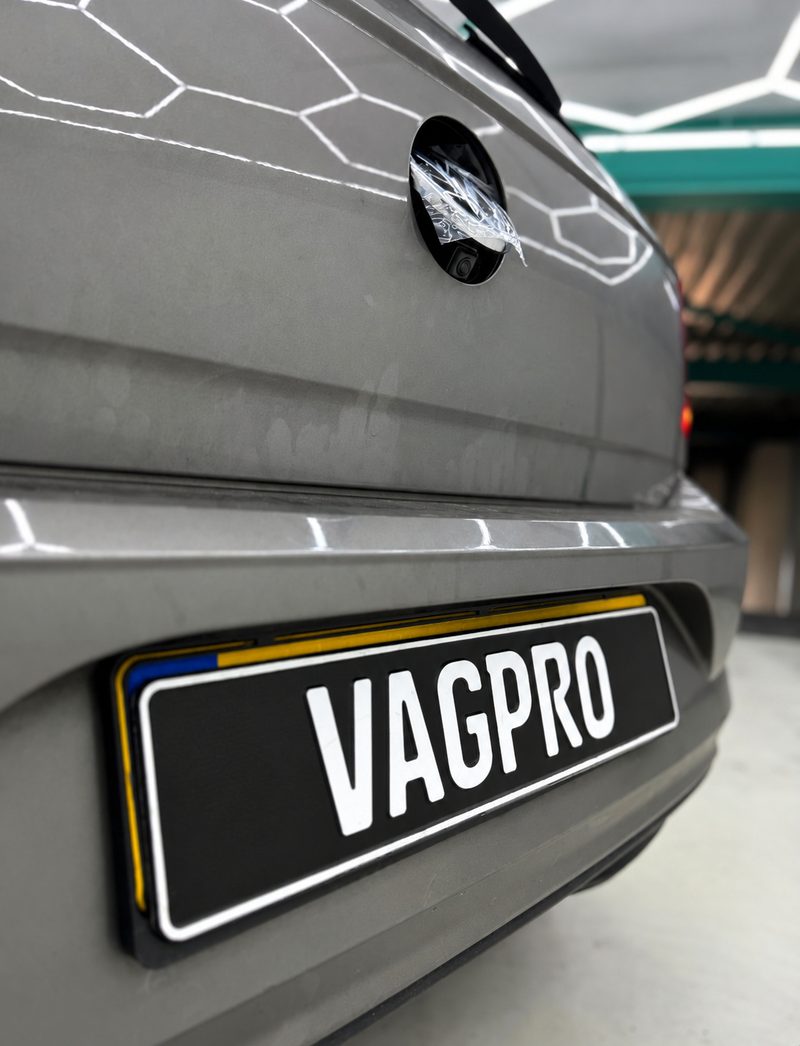 Grijze Volkswagen met VAGPRO-kentekenplaat. Achter het opengeklapte VW-embleem zit de achteruitrijcamera, met de beschermfolie er nog op